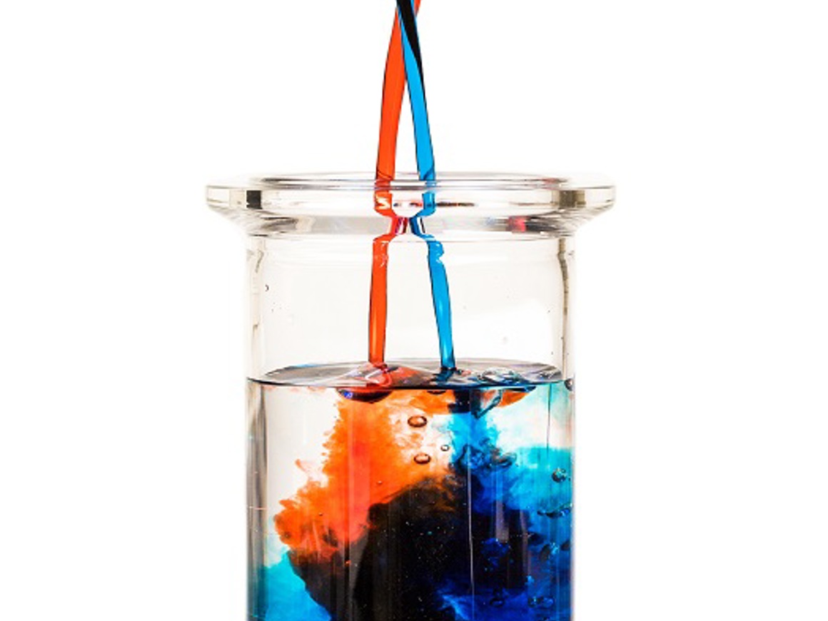A glass beaker containing clear liquid where orange and blue dyes are being separately introduced through two tubes, creating distinct streams of color in the solution. The dyes maintain separation, demonstrating selective extraction.